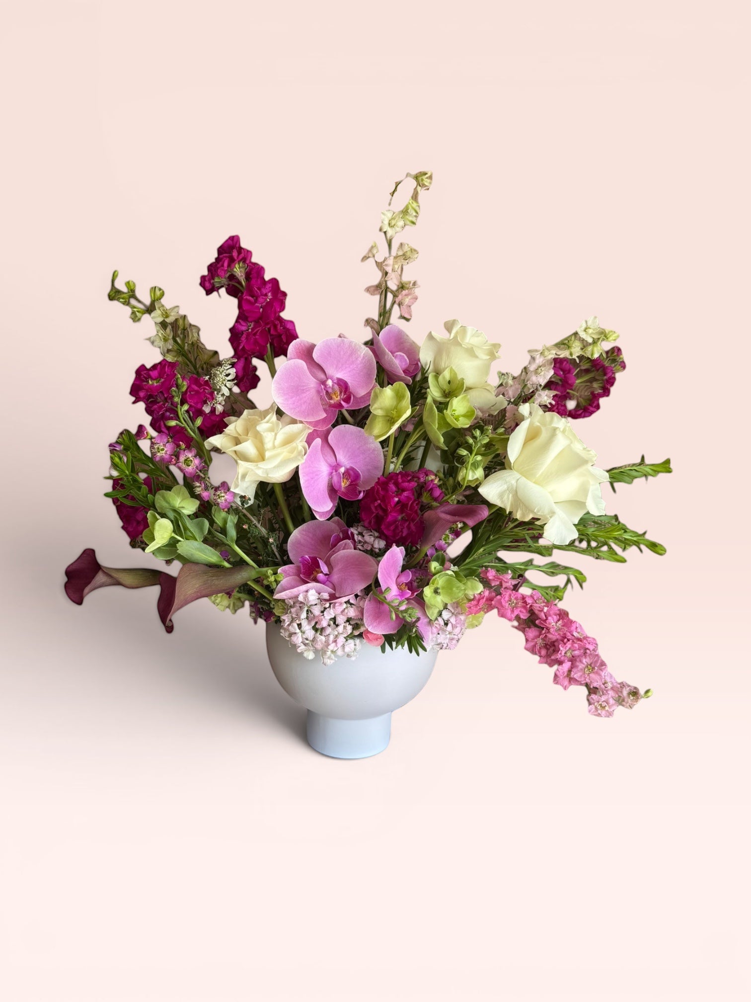 Skye Rose Floral Plum Essence arrangement featuring pink orchids, white roses, and plum calla lilies in a white vase.