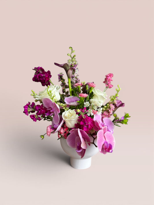 Skye Rose Floral Plum Essence arrangement featuring purple orchids, calla lilies, and white roses in a white pedestal vase